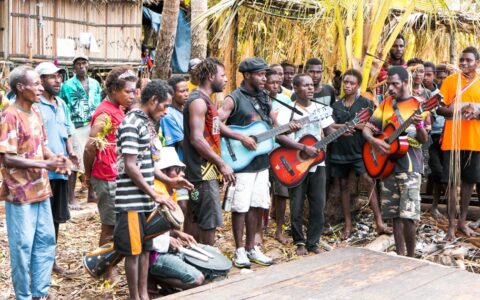 Local men stand and play drums & guitars while singing among thatch buildings, seen on a Papua New Guinea cruise.