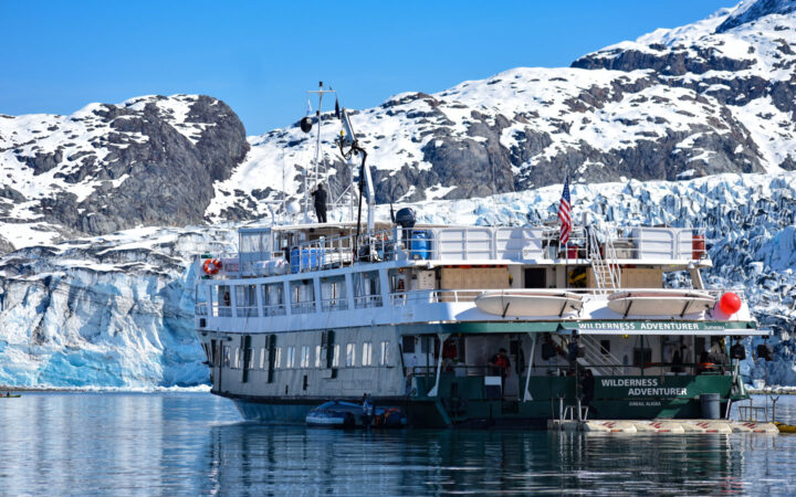 White & green small ship Wilderness Adventure floats close in front of a massive icy glacier on a sunny day.