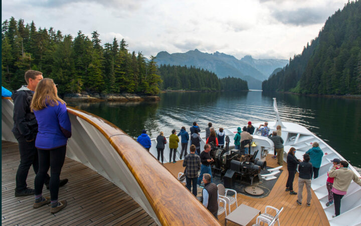 Guests stand on decks of small ship while sailing calm inlet on the Glacier Bay National Park with Inside Passage & San Juan Islands cruise.