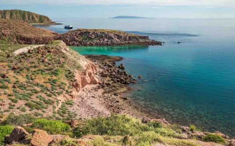 Aerial view of Baja shoreline with sand, dusty rock, green shrubs & turquoise sea with small ship sitting in the distance.