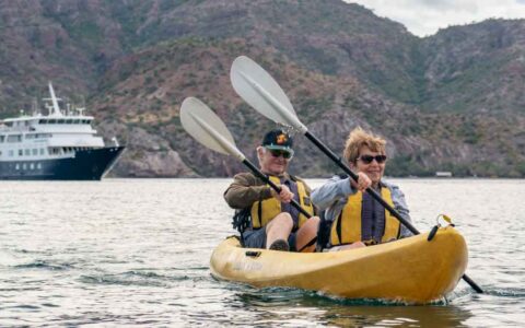 Man & woman paddle a yellow tandem kayak on calm water with a small blue & white ship & rocky hillside behind in Baja.