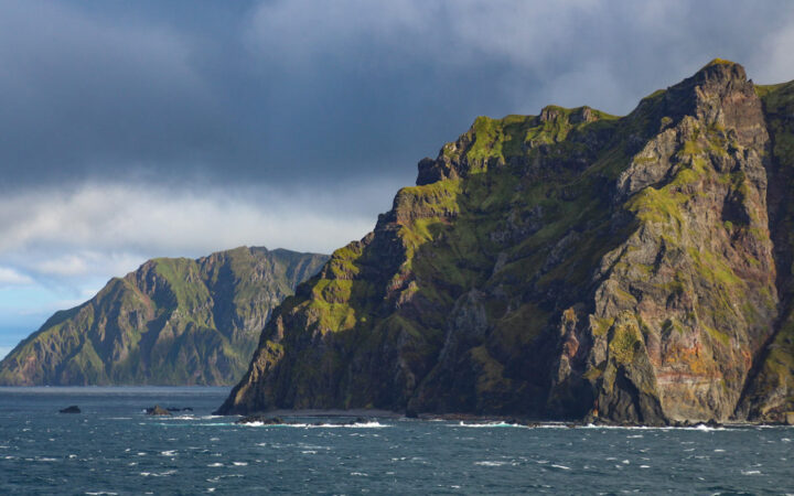 Jagged volcanic mountains of rock with green moss rise from dark blue seas in the Aleutian Islands, Alaska. on a cloudy day.