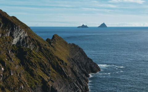 Kerry cliffs of green & dark rock drop sharply into blue ocean with smaller islands offshore on a sunny day in Ireland.