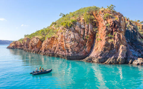 Zodiac of people pulls up to rocky red islet with teal-colored water & bright green plants in Australia's Kimberley region.
