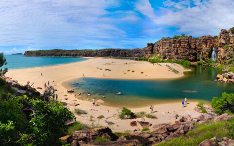 Aerial view of Tranquil Bay, Australia, with white-sand spit, turquoise water, red-rock cliffs, waterfalls & lush greenery.