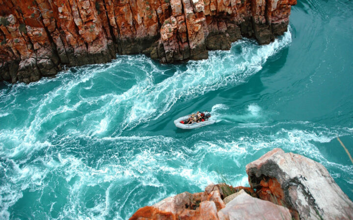 A Zodiac boat takes travelers against the current of a turquoise river between red cliffs on The Kimberley Cruise in Australia.