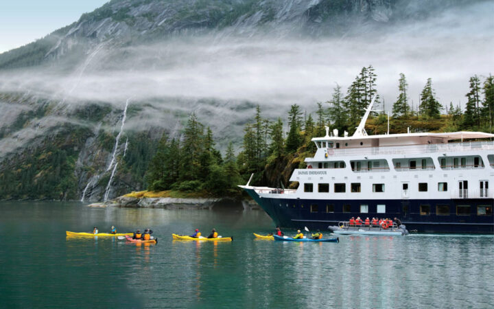 In front of tall fjord walls with waterfalls the small Safari Endeavour ship floats as a group of kayakers paddle.