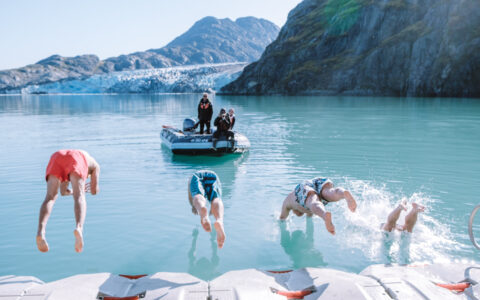 Guests leaping off the kayak towards zodiac platform into glacier bay for a polar plunge on a bright sunny day