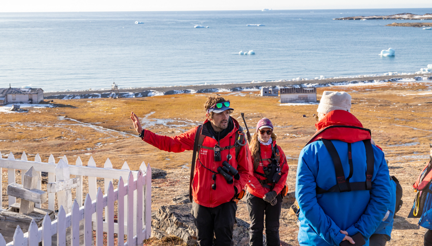 Guide holds his hand out to point out old grave site off the shores of the Arctic next to small shacks on a sunny day