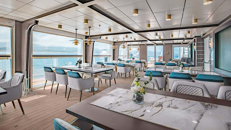 Seating aboard small ship Ocean Victory cruise with large floor-to-ceiling windows, tables of 6 set for guests to enjoy