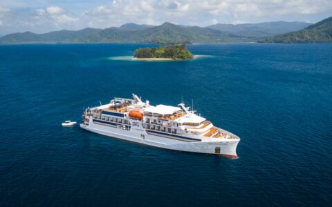 Aerial view of a small white expedition ship cruising a Papua New Guinea cruise in deep blue water beside green islands.