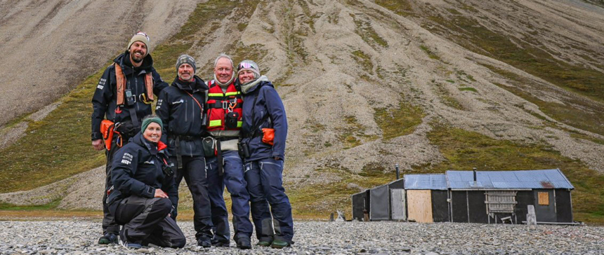 Polar Quest Svalbard guides in black, blue & red jackets pose in front of a historic trapper's hut backed by green hillside.
