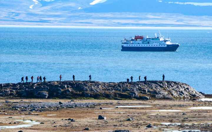 Small group of Polar Quest Svalbard travelers hike a rocky ridgeline among golden tundra backed by teal sea & a small ship.