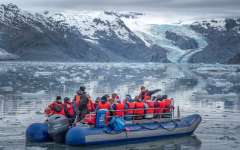 Explorers with red lifevests navigating through the icy still waters next to the run off of a large glacier off in the distance
