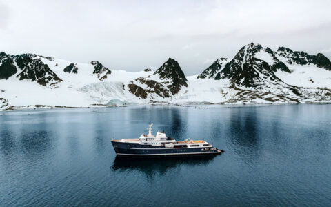 Superyacht Aqua Lares with dark blue hull & multiple decks sits in calm sea beside snowy peaks on a cloudy day in Svalbard.