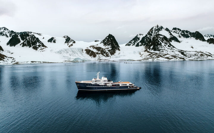 Superyacht Aqua Lares with dark blue hull & multiple decks sits in calm sea beside snowy peaks on a cloudy day in Svalbard.