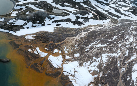Aerial view of small ship sitting in bay beside snowy peaks, teal sea & rust-colored outwash in Svalbard, the Arctic.