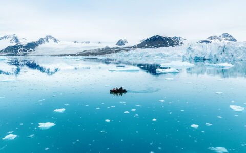 Aerial view of a Zodiac small tender vessel cruising in icy teal sea beside a glacier & snow-covered peaks in Svalbard.