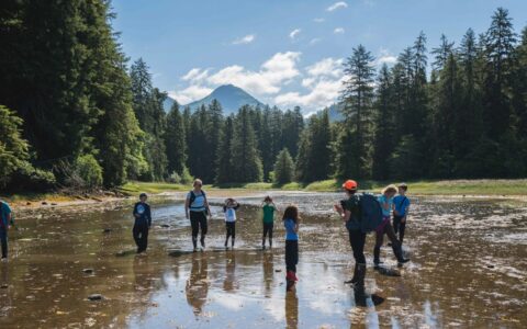 Group of kids & 2 adults hike through a shallow, muddy tidal outwash surrounded by forest on a sunny day during an Alaska family cruise.
