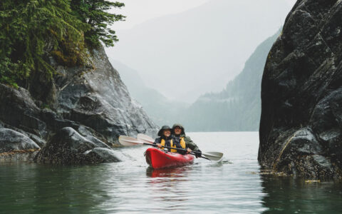 Tandem kayakers in a red boat paddle calm water between large dark boulders on a misty day during the Wild & Wooly Alaska Cruise.