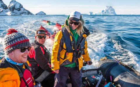 Antarctic travelers Zodiac cruising with guide standing and steering the inflatable boat & mountains in the background on a sunny day.