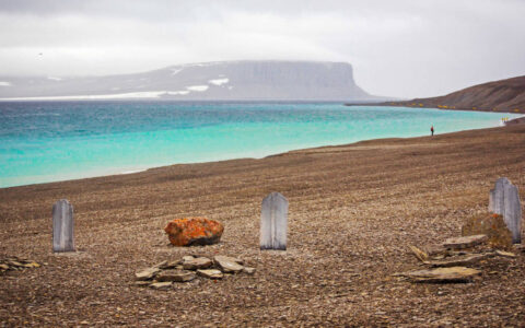 Beechey Island with 3 graves of explorers on a misty day with turquoise water along the brown, rocky shoreline, seen during The Northwest Passage Canadian High Arctic voyage.