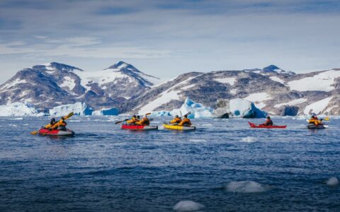 5 tandem paddlers travel on icy blue waters with snowy mountains in the background during The Northwest Passage Canadian High Arctic voyage.