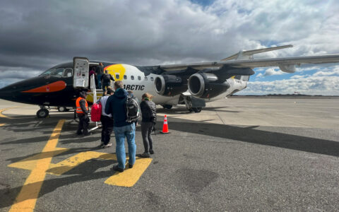 Small line of Active Antarctica air cruise travelers stands on tarmac waiting to board a small jet painted like a penguin.