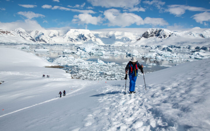 Active & Wild Antarctica air cruise travelers hike over snow with iceberg-filled bay behind on a sunny day.