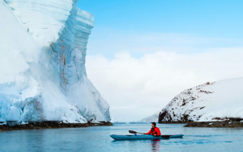 Man in bright red jacket paddles blue kayak in calm sea past towering glacier of snow & ice on a blue-sky day in Antarctica.