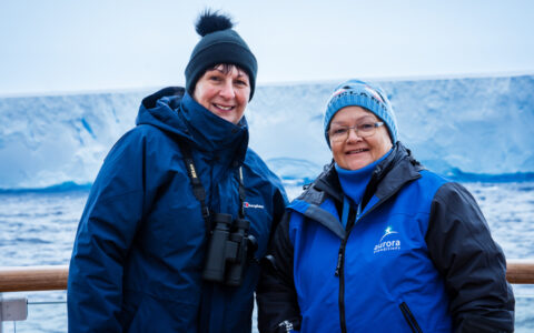 2 women in blue jackets & knit hats stand on deck of small expedition ship with massive glacier behind them.