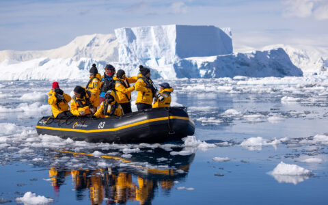 Antarctica travelers cruise in a black Zodiac boat among glassy sea with iceberg bits near a snowy glacier.