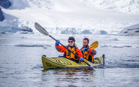 Travelers paddle yellow kayak through cold water by snowfields on a sunny day in Antarctica after flying the Drake Passage.