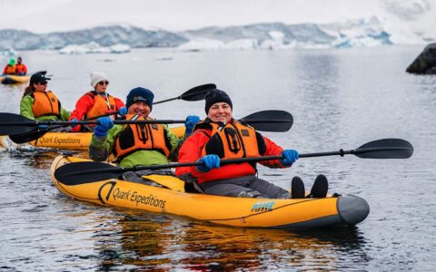 Polar travelers pose for the camera while paddling an inflatable yellow kayak among snowfields in calm water.