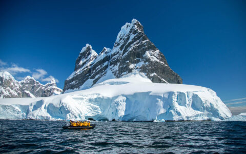 Polar travelers in yellow jackets cruise calm water in a black Zodiac boat beside a snowfield under blue sky in Antarctica.