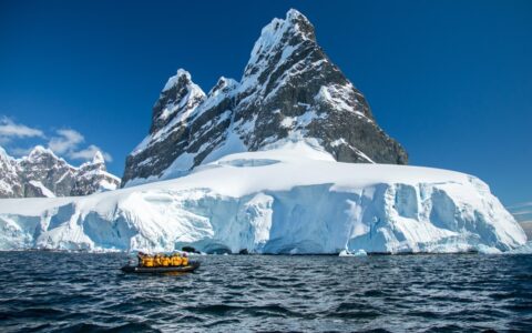 Polar travelers in yellow jackets cruise calm water in a black Zodiac boat beside a snowfield under blue sky in Antarctica.