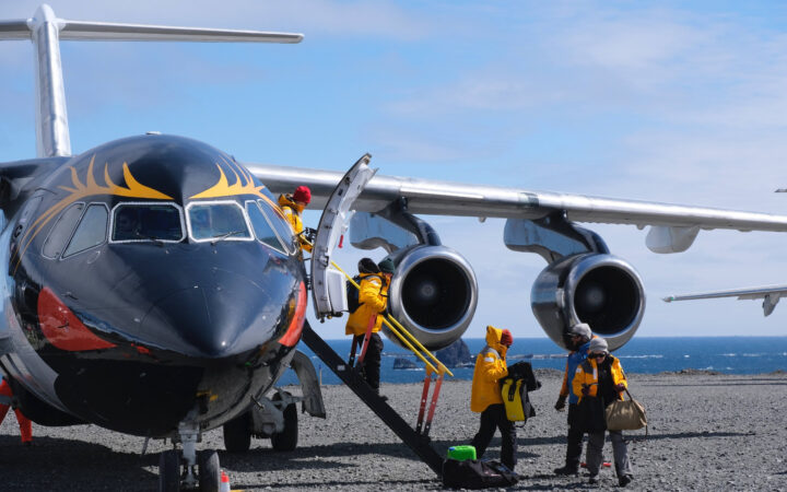 Antarctic Express travelers board a small jet painted like a macaroni penguin with yellow eyebrows & orange cheeks on a sunny day.