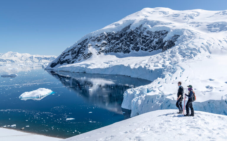 2 Antarctica air Cruise travelers stand in snowshoes atop a snowy peak overlooking a glassy bay with icebergs in the sun.