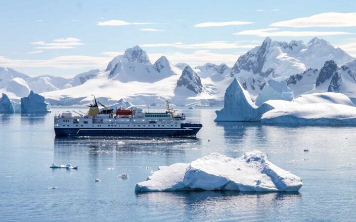 A small ship cruises Antarctica among giant white icebergs and snowcapped mountains.