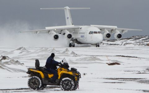 A man rides a yellow all terrain vehicle across Antarctic tundra, beyond him a white charter plane sits on a landing strip in Antarctica.