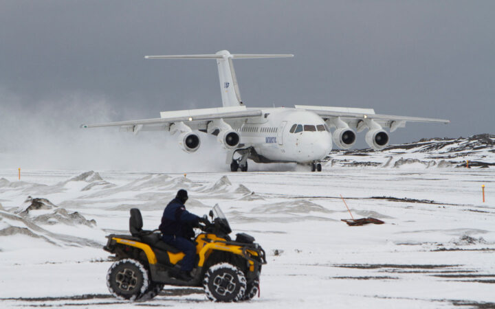 A man rides a yellow all terrain vehicle across Antarctic tundra, beyond him a white charter plane sits on a landing strip in Antarctica.
