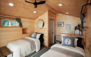 Wood cabin with two twin beds, tan bedding and grey pillows, with a fan and small windows