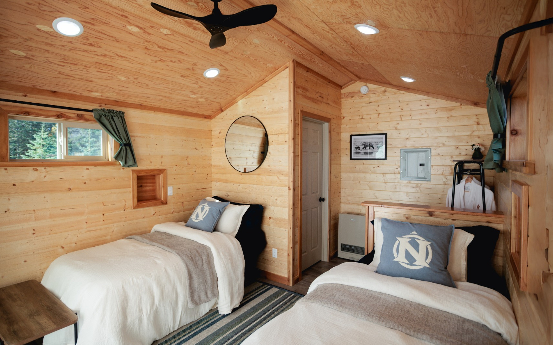 Wood cabin with two twin beds, tan bedding and grey pillows, with a fan and small windows