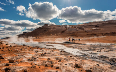 Iceland travelers walk over Hverarondor Hverir geothermal area with its multicolored, cracked mud & steam under clouds.