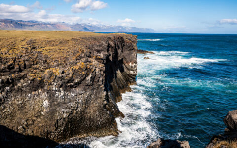 Scenic Iceland coast with golden tundra, tan cliffs & turquoise sea with large swells hitting the shore on a sunny day.