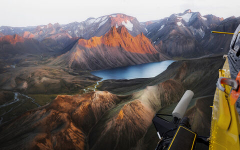 View from yellow helicopter as it flies over jagged peaks with green valleys & a small lake in Iceland.