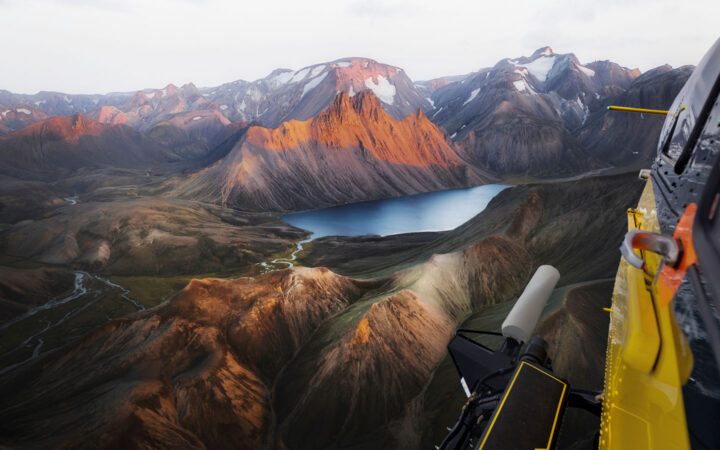 View from yellow helicopter as it flies over jagged peaks with green valleys & a small lake in Iceland.