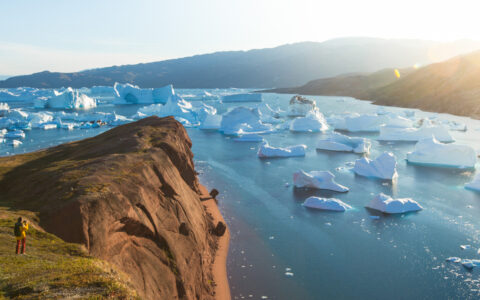 A hiker looks our over a bay full of icebergs on a bright day while exploring Iceland & Greenland on a trip Under the Northern Lights.