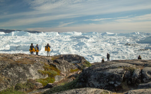 Hikers on a Northwest Passage small ship cruise stand at an overlook of tundra & rock looking onto an ice sheet in the sun.