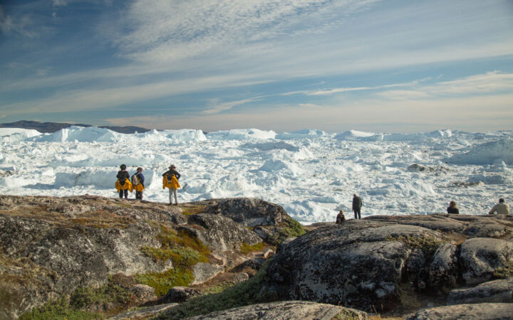 Hikers on a Northwest Passage small ship cruise stand at an overlook of tundra & rock looking onto an ice sheet in the sun.